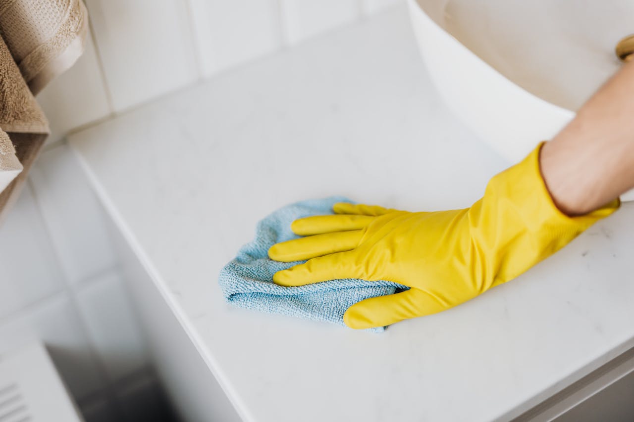 our-story From above crop unrecognizable person with microfiber cloth wearing yellow rubber glove and cleaning white marble tabletop of vanity table with washbasin in bathroom